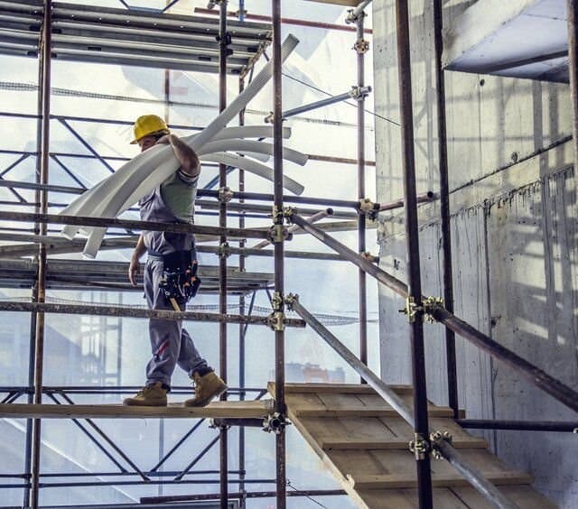 A worker in a yellow helmet carrying scaffold tubes along a boarded scaffold platform beside a building under construction.