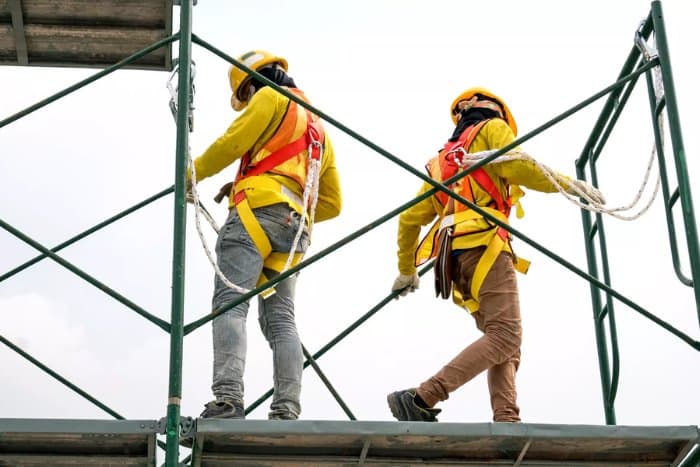 Two scaffolders in yellow jackets, helmets and full-body harnesses with lanyards working on a boarded scaffold platform against a bright sky.