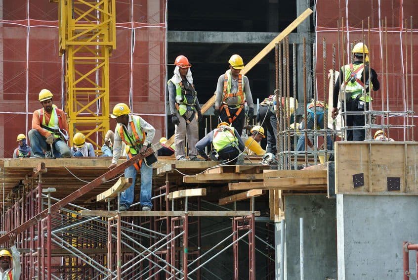 Scaffolders working from a timber deck on a concrete frame, with a tower crane and reinforcement steel behind them.