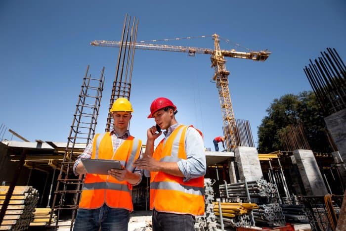Two site engineers in hi-vis vests, one checking a tablet and one on a phone call, in front of a tower crane and reinforcement cages.