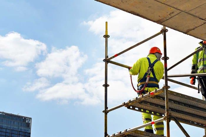 Scaffolder in a hi-vis jacket and helmet, clipped on with a fall-arrest lanyard, standing on a scaffold platform against a clear sky.