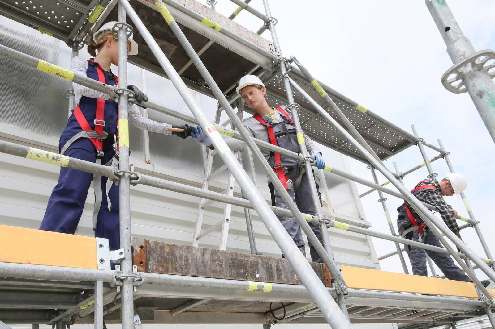 Three scaffolders in helmets and harnesses assembling a galvanised steel system scaffold with timber platform boards on a construction site.