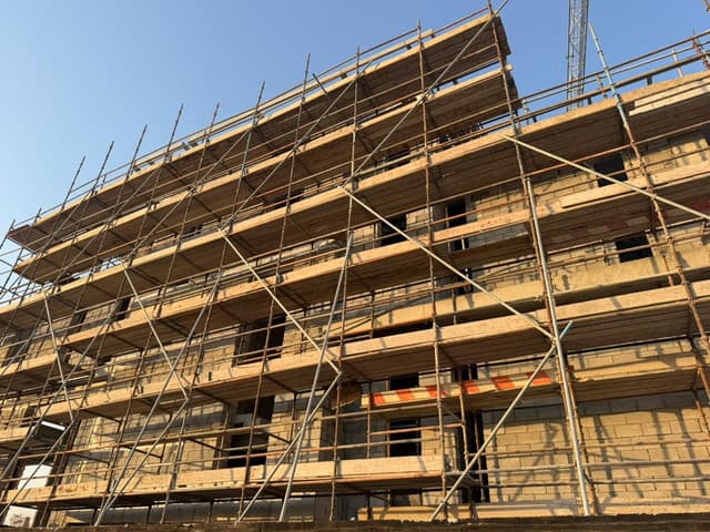 Multi-lift tube-and-fitting scaffold with timber platform boards and diagonal bracing across the blockwork facade of a building under construction.