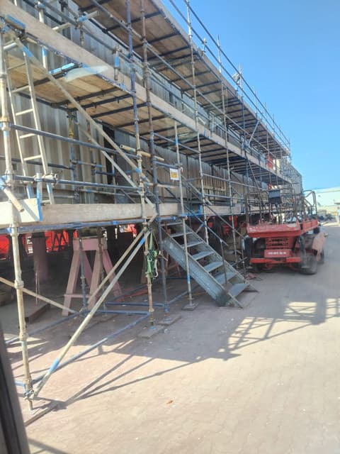 Tube-and-fitting scaffold with timber platform boards, blue ladders and a stair unit erected along the facade of a building, with a scissor lift parked beside it.