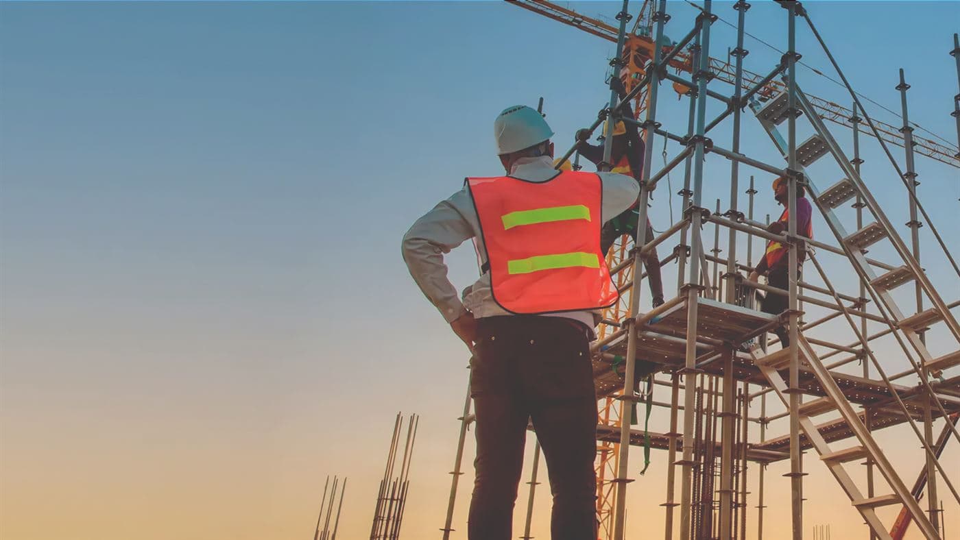 A frame scaffold standing beside a structure under construction, with workers on the upper platform and a tower crane behind.