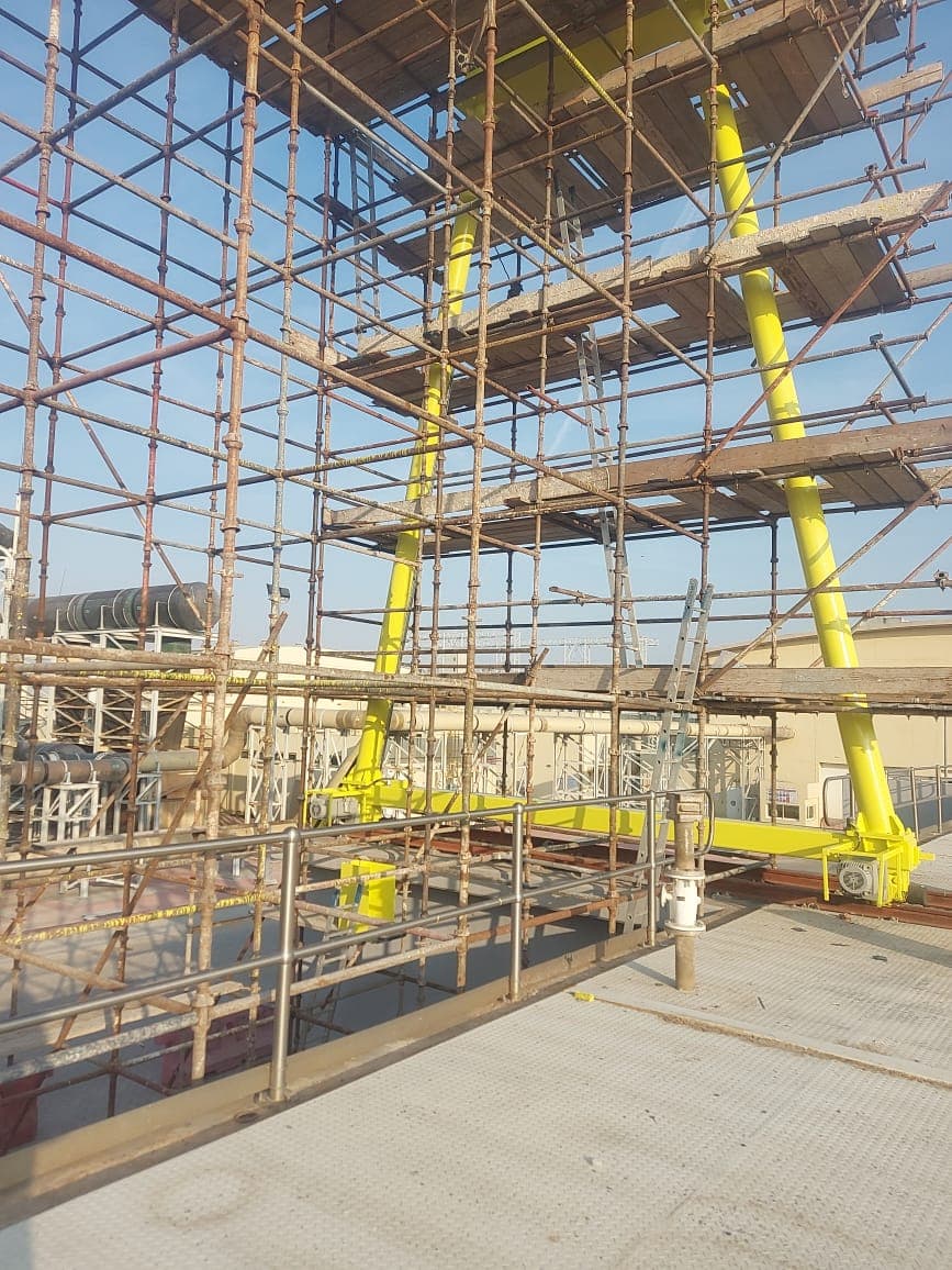 Tube-and-fitting scaffold with timber platforms and ladders erected around pipework and steelwork inside an industrial plant, with yellow-painted members running through the structure.