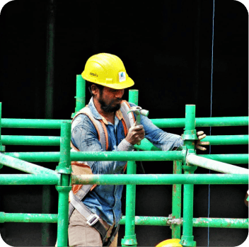 Scaffolder in a yellow helmet and an orange high-visibility vest working on green-painted scaffold tubes with a hand wrench.