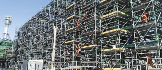 Multi-level access scaffold with yellow platform boards and guard rails covering the face of a refinery plant, with workers in orange visible on several lifts.