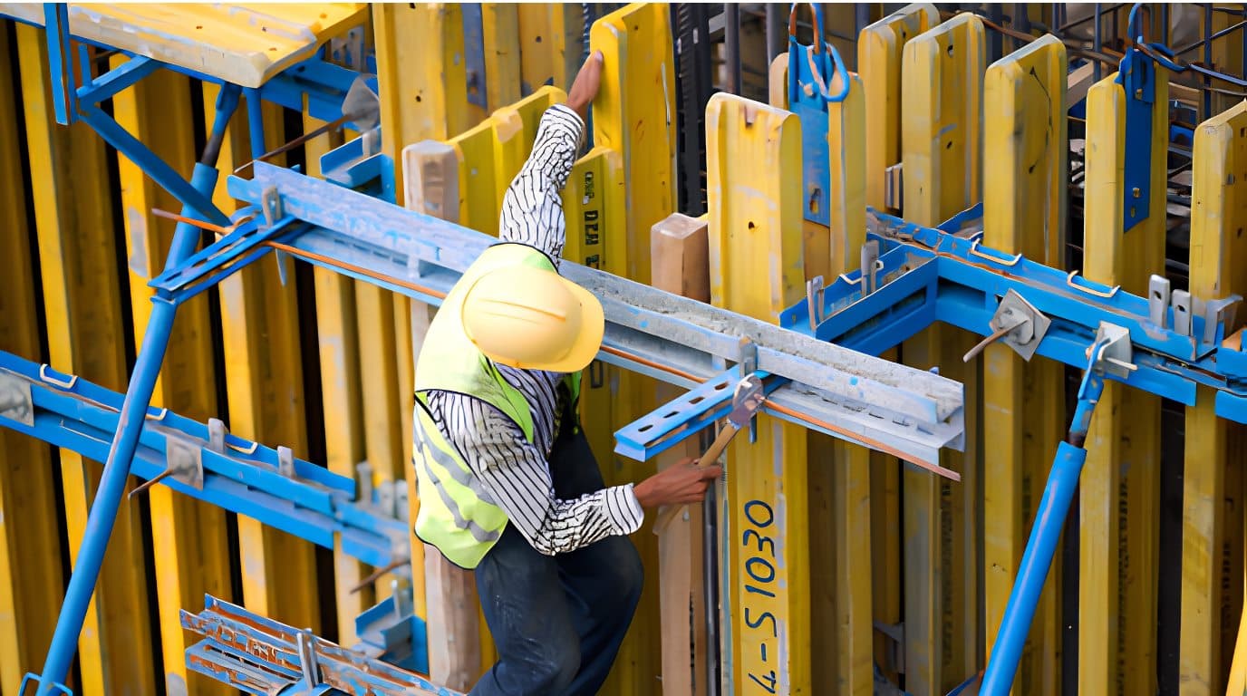 Worker in a hi-vis vest and helmet fixing a steel waler across yellow formwork panels on a concrete wall.