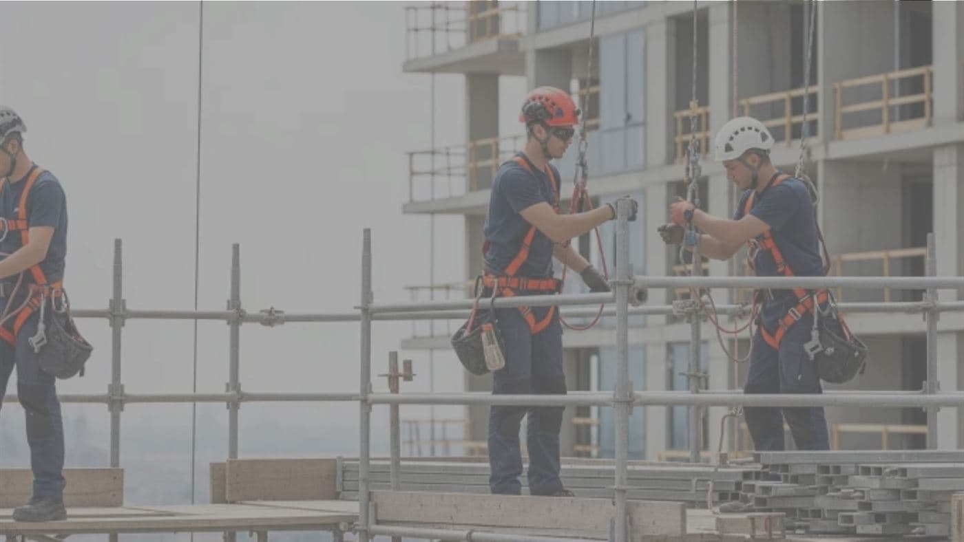Workers wearing full-body fall-arrest harnesses, lanyards and helmets, standing and working on a scaffold platform in front of a building under construction.