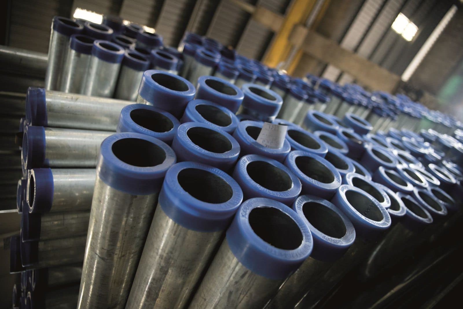 Stacked hot-dip galvanised steel pipes and tubes with blue protective end caps inside a warehouse.