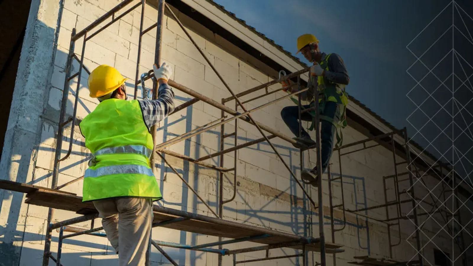 Two workers in high-visibility vests, helmets and harnesses assembling a tube-and-fitting scaffold against a blockwork wall in low sunlight.