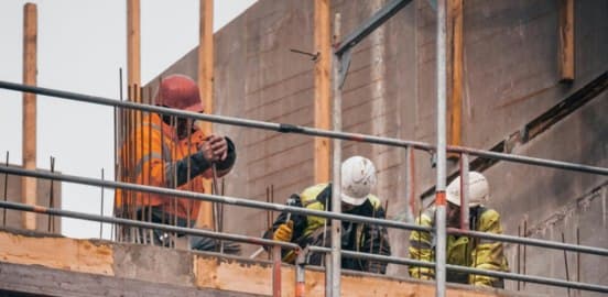 Three workers in high-visibility clothing and helmets working from a scaffold deck on a concrete structure, with formwork and reinforcement behind them.