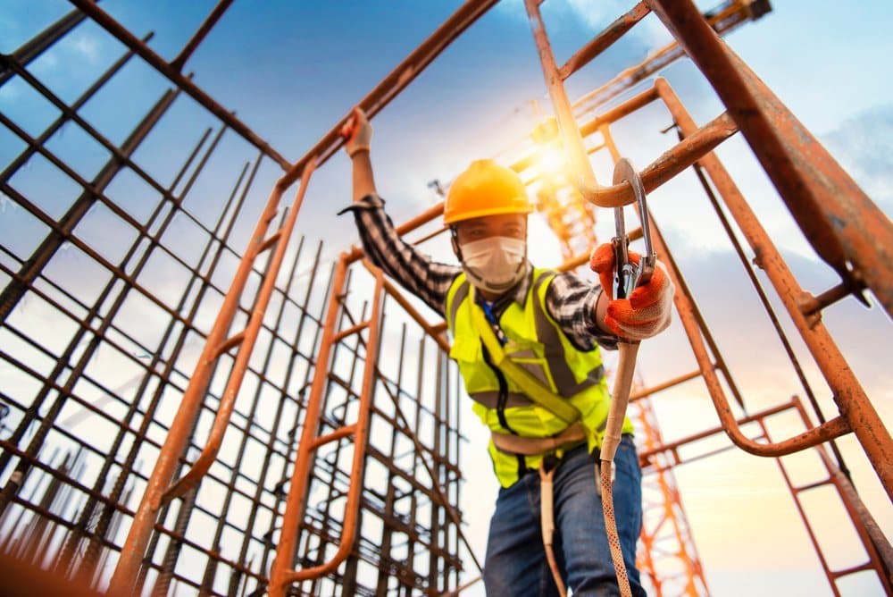 Worker in a yellow helmet, face mask and hi-vis vest wearing a safety harness and holding a lanyard hook beside orange-painted steel frames and reinforcement bars.